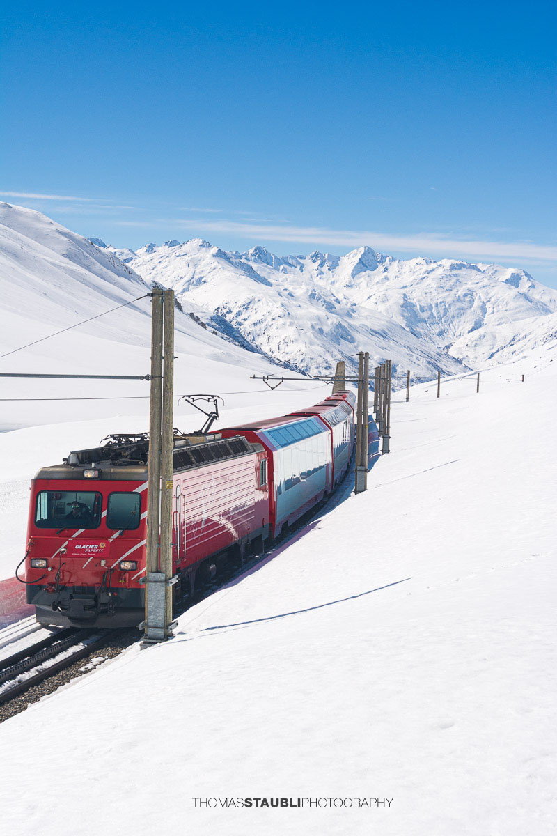 Glacier Express fährt durch die verschneite Landschaft am Oberalppass mit Blick ins Urserental.