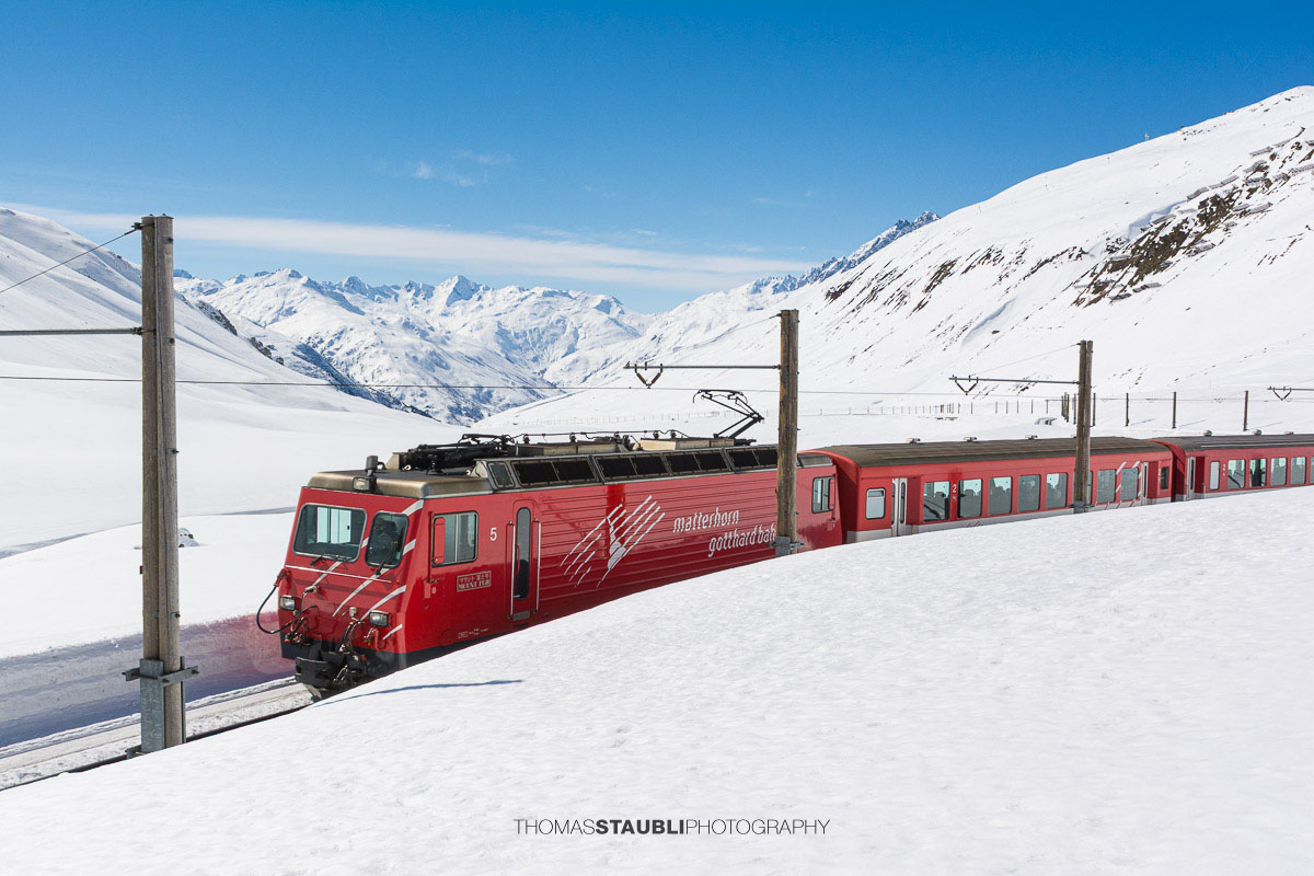 Matterhorn Gotthard Bahn fährt durch die verschneite Landschaft am Oberalppass mit Blick über das Urserental.