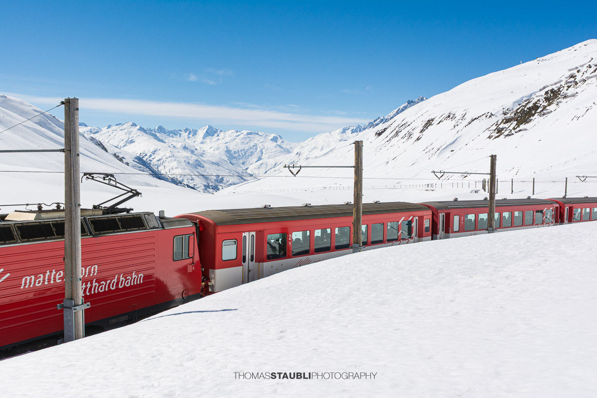 Matterhorn Gotthard Bahn fährt durch die verschneite Landschaft am Oberalppass mit Blick über das Urserental.