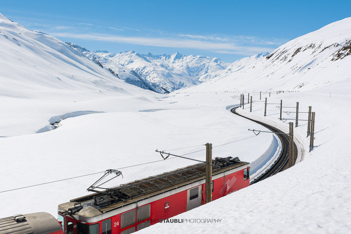Matterhorn Gotthard Bahn fährt durch die verschneite Landschaft am Oberalppass mit Blick über das Urserental.