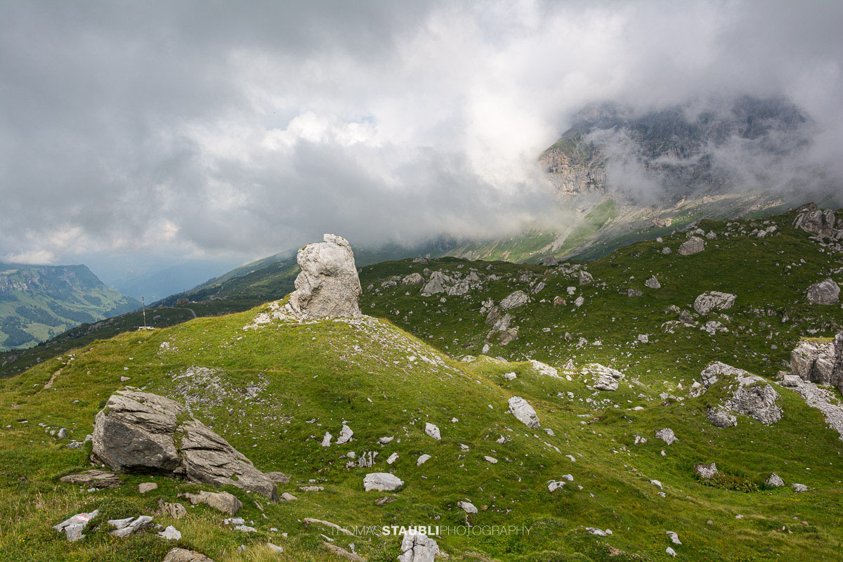 Aufziehende Wolken und Nebel über dem Griessbödemli am Klausenpass mit markantem Felsblock im Vordergrund.