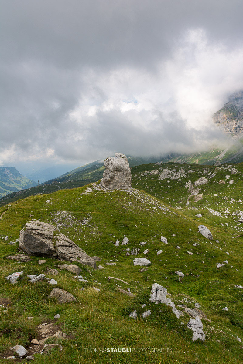 Aufziehende Wolken und Nebel über dem Griessbödemli am Klausenpass mit markantem Felsblock im Vordergrund.