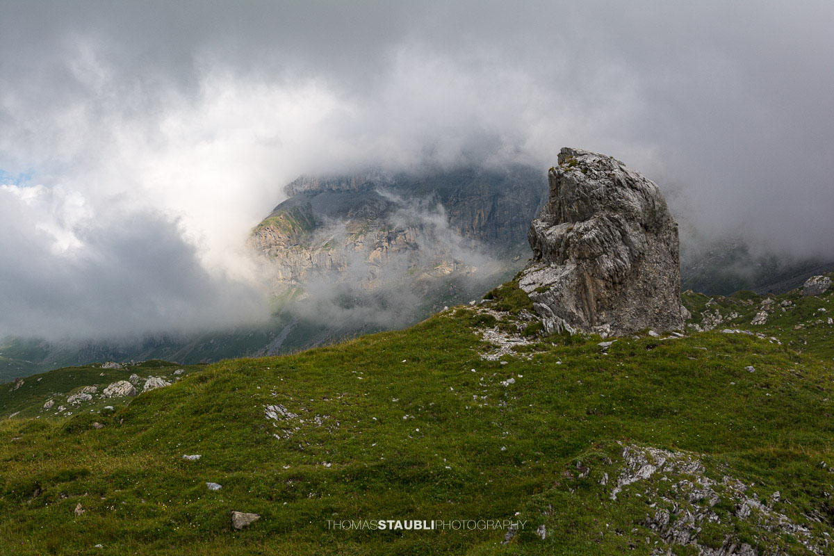 Aufziehende Wolken und Nebel über dem Griessbödemli am Klausenpass mit markantem Felsblock im Vordergrund.