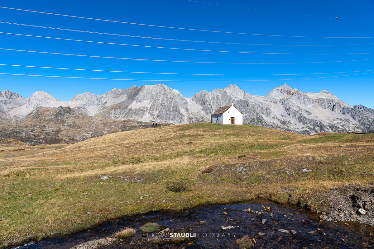 Die kleine Kapelle San Giacomo im Bedrettotal vor der Kulisse hoher Berge, mit einem Bergbach im Vordergrund.