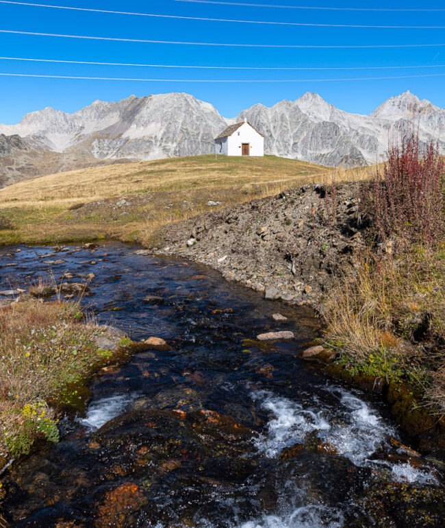 Die kleine Kapelle San Giacomo im Bedrettotal vor der Kulisse hoher Berge, mit einem Bergbach im Vordergrund.