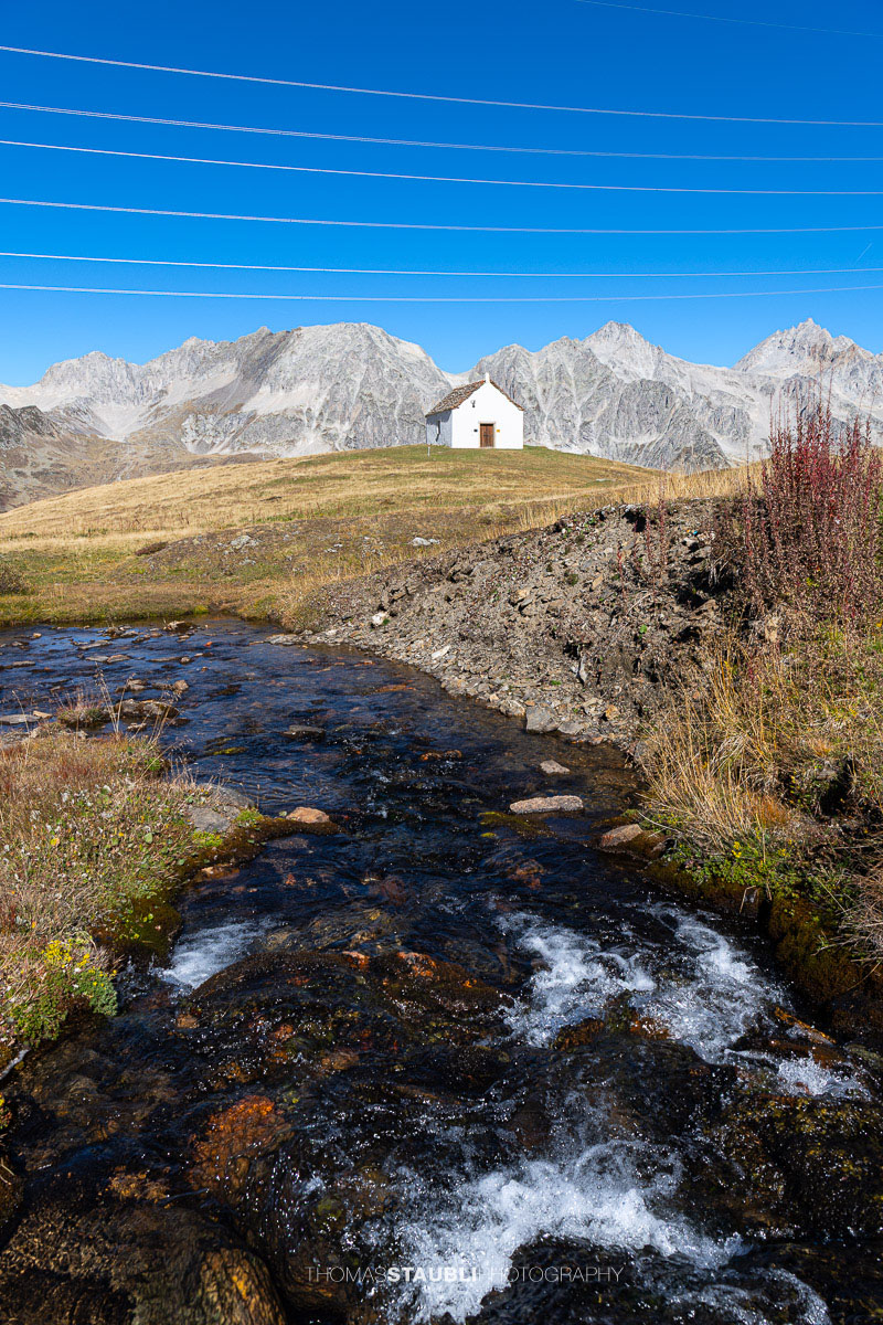 Die kleine Kapelle San Giacomo im Bedrettotal vor der Kulisse hoher Berge, mit einem Bergbach im Vordergrund.